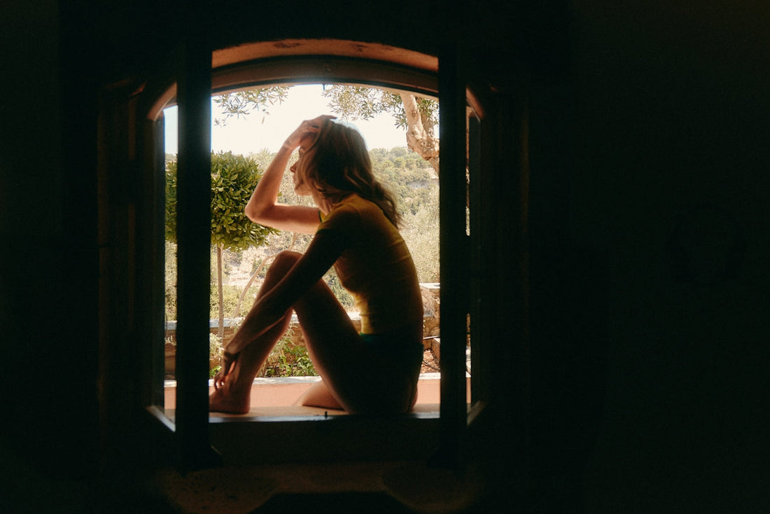 Woman in a soft yellow top and underwear sitting on a sunny windowsill, looking out at trees