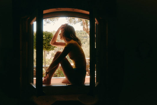 Woman in a soft yellow top and underwear sitting on a sunny windowsill, looking out at trees